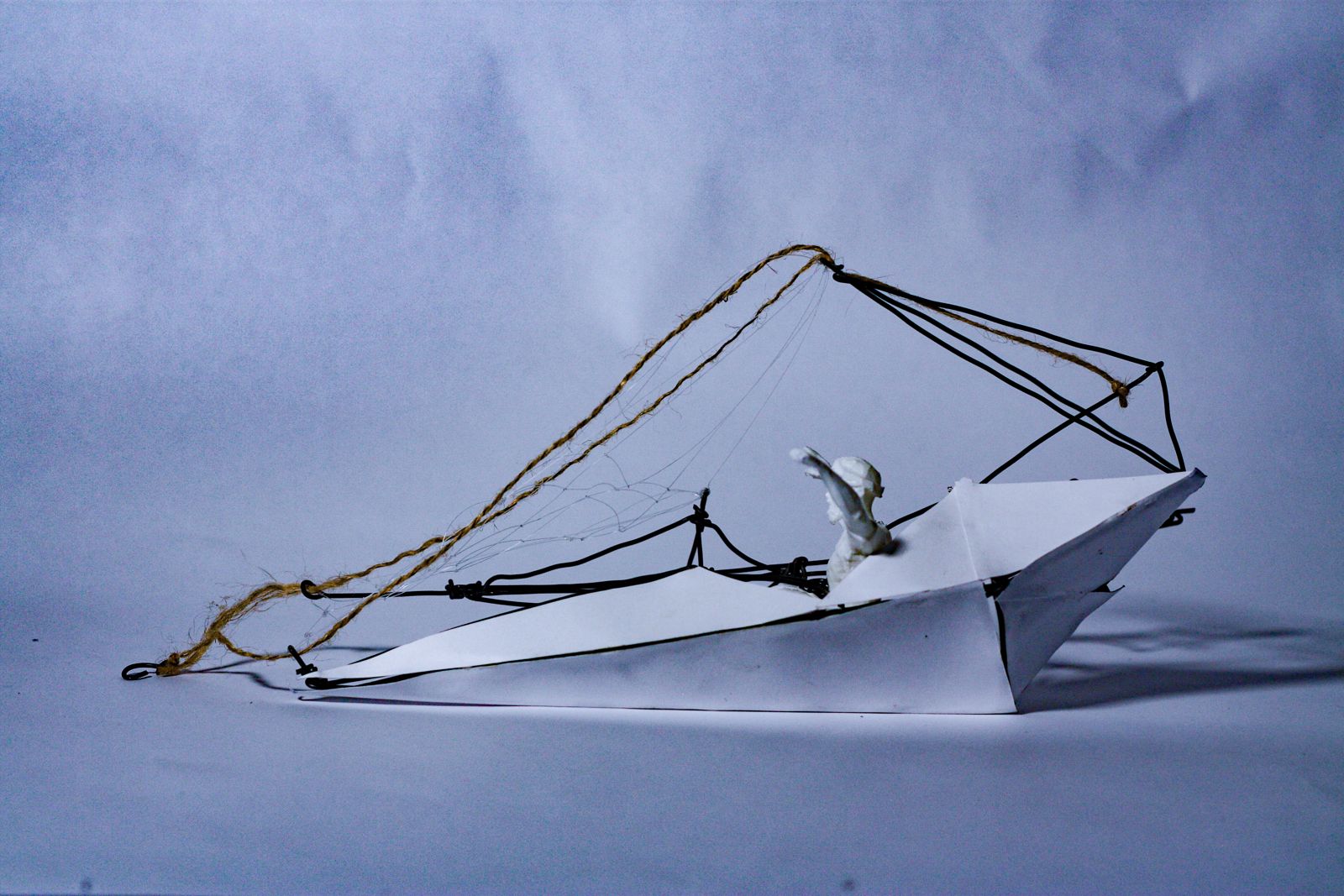 A small boat model on a pale grey backdrop: a folded white paper hull, a black wire frame rising over the stern, fine netting strung between the wires, and a length of tan twine trailing to a hook at the bow. A tiny white figure sits in the middle.