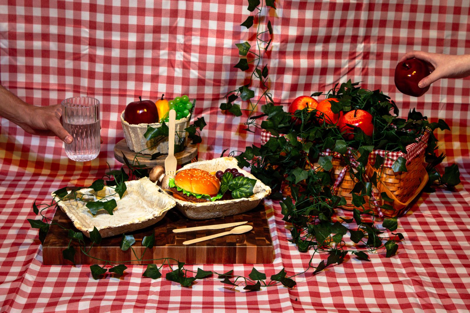 A picnic on red gingham: a mycelium takeout box holding a burger, a mycelium bowl of fruit, wooden utensils and ivy, with hands reaching in for a glass and an apple.